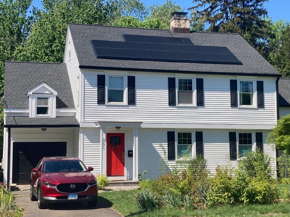 A red car is parked in front of a white house with solar panels on the roof