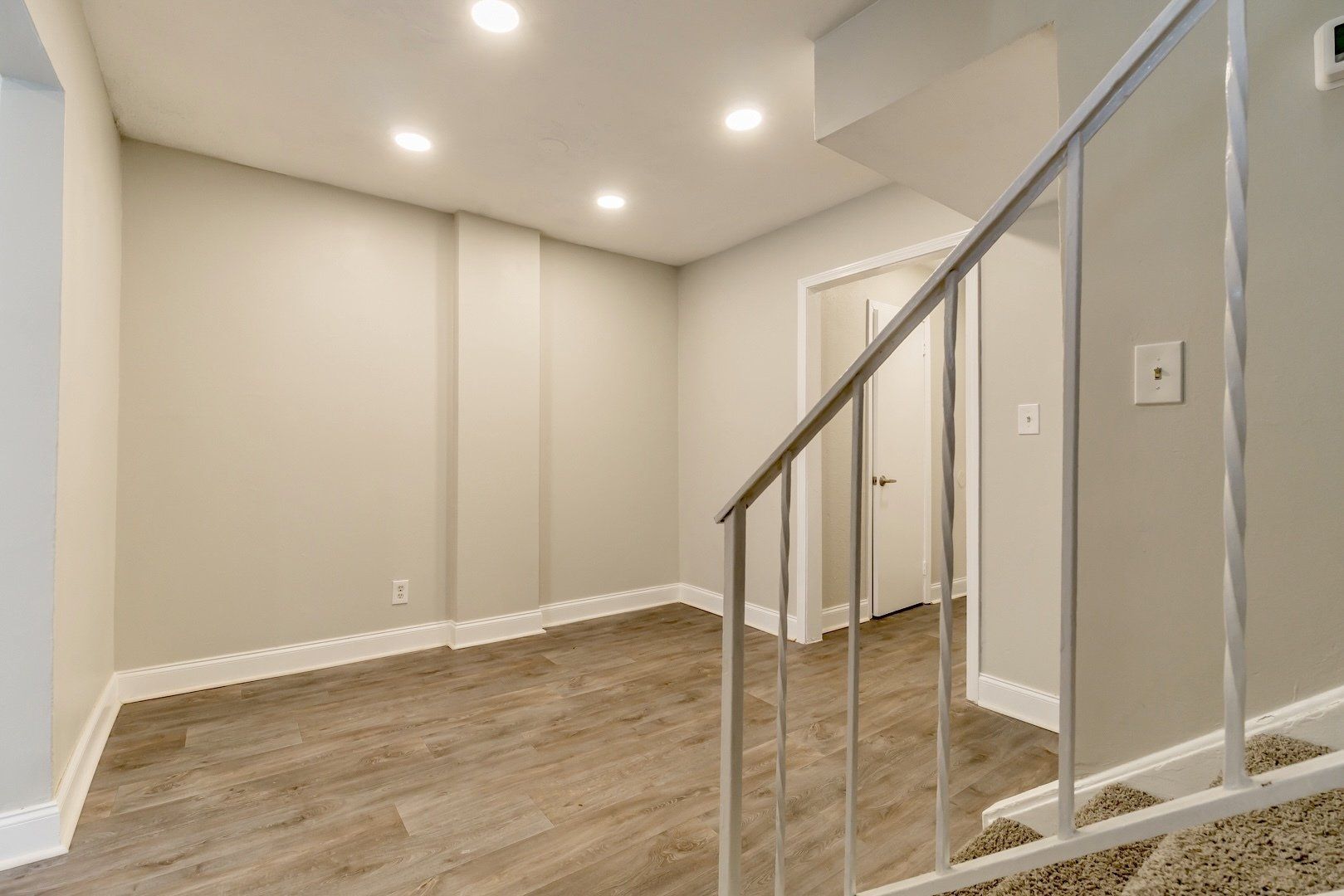 Empty beige room with wood flooring, recessed lights, and a stair railing in the foreground