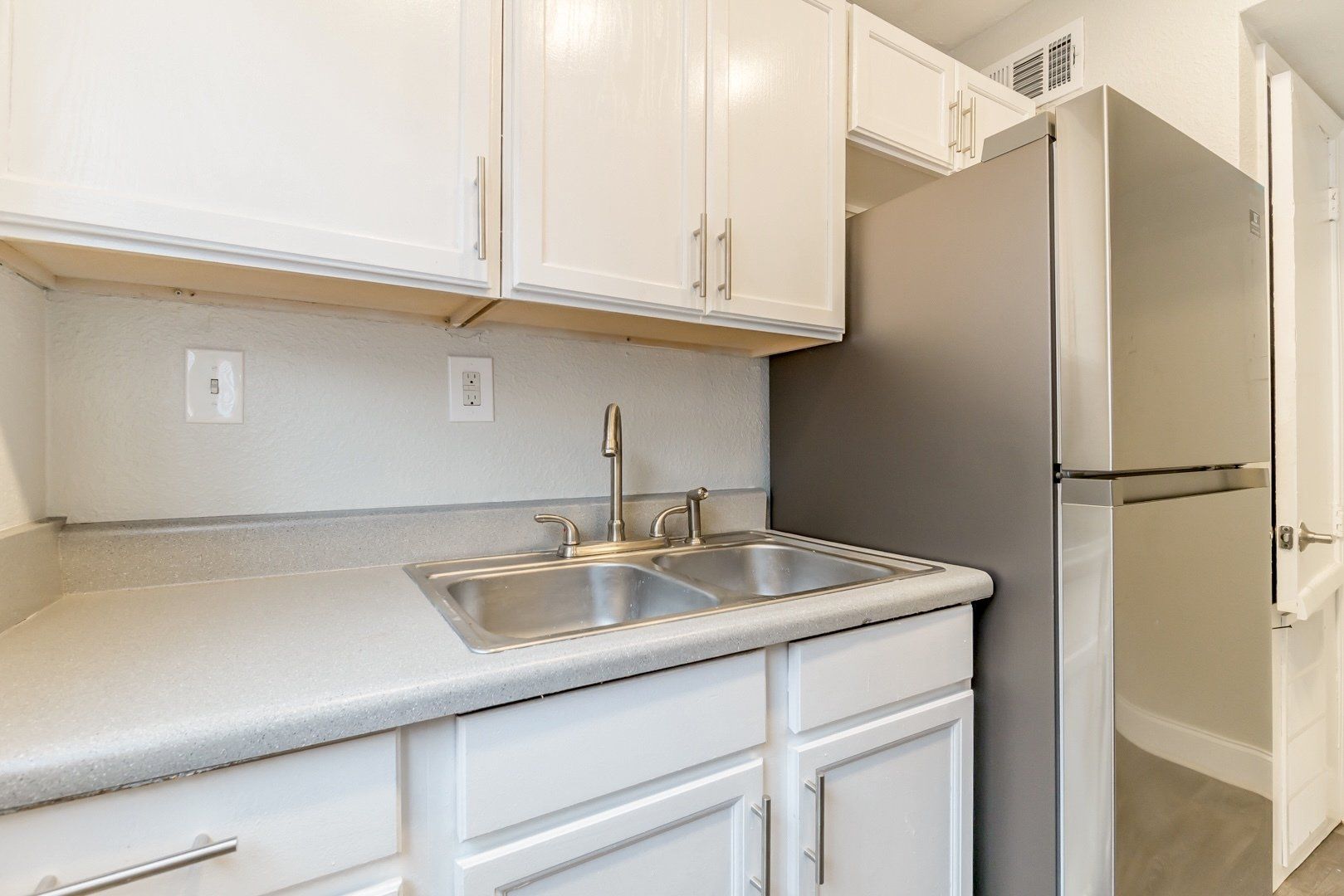 Small kitchen corner with white cabinets, double sink, and stainless steel refrigerator.