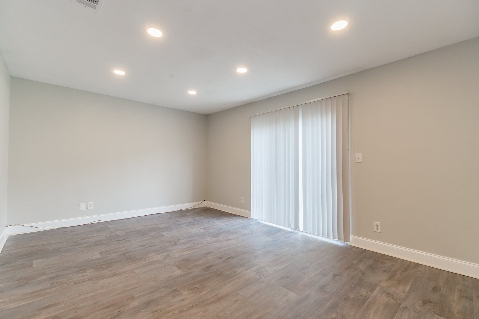 Empty beige living room with wood floors, recessed lights, and sliding glass door with vertical blinds