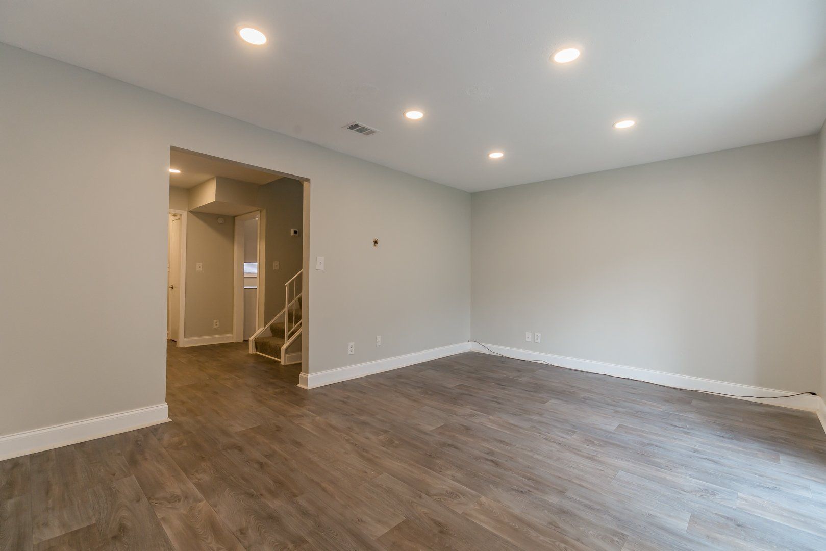 Empty living room with gray walls, wood floors, recessed lights, and a hallway opening to stairs.