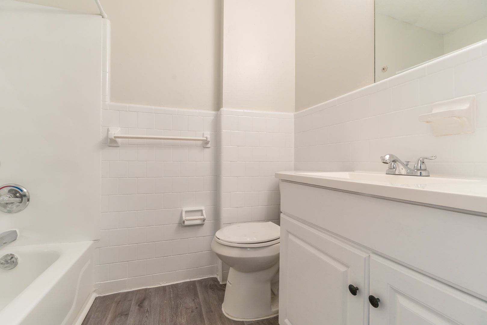 Small white bathroom with tub, toilet, sink vanity, towel rack, and wood-look flooring.