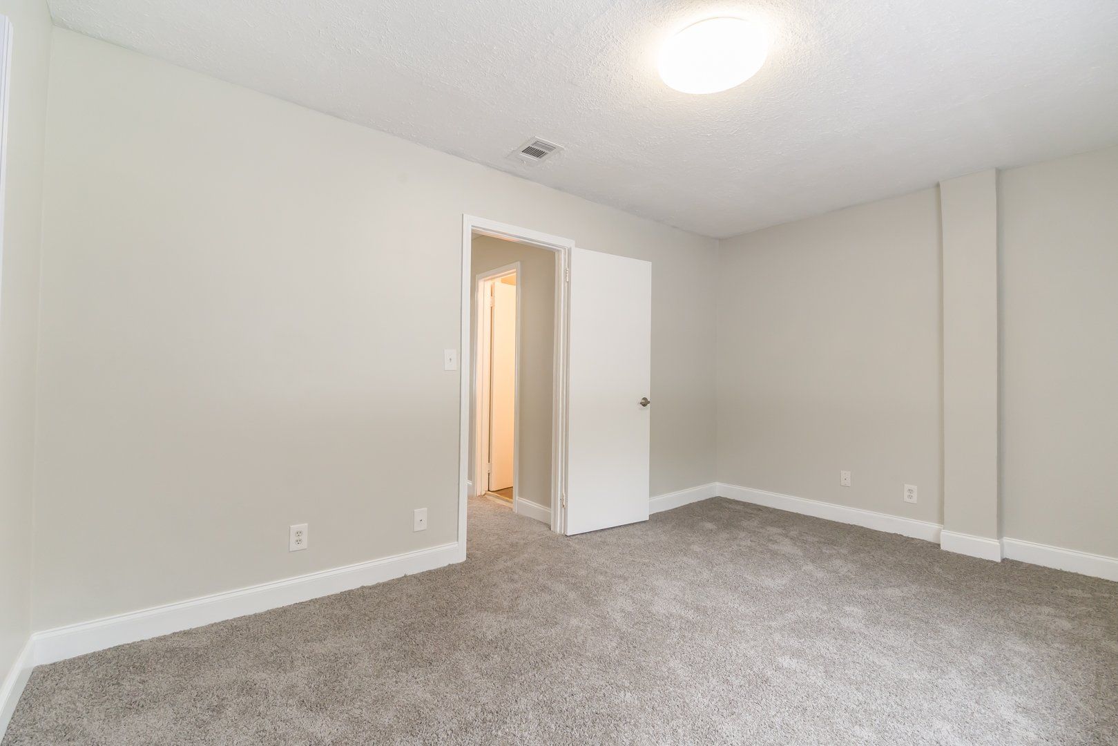 Empty beige bedroom with carpet, ceiling light, and open door to a lit hallway