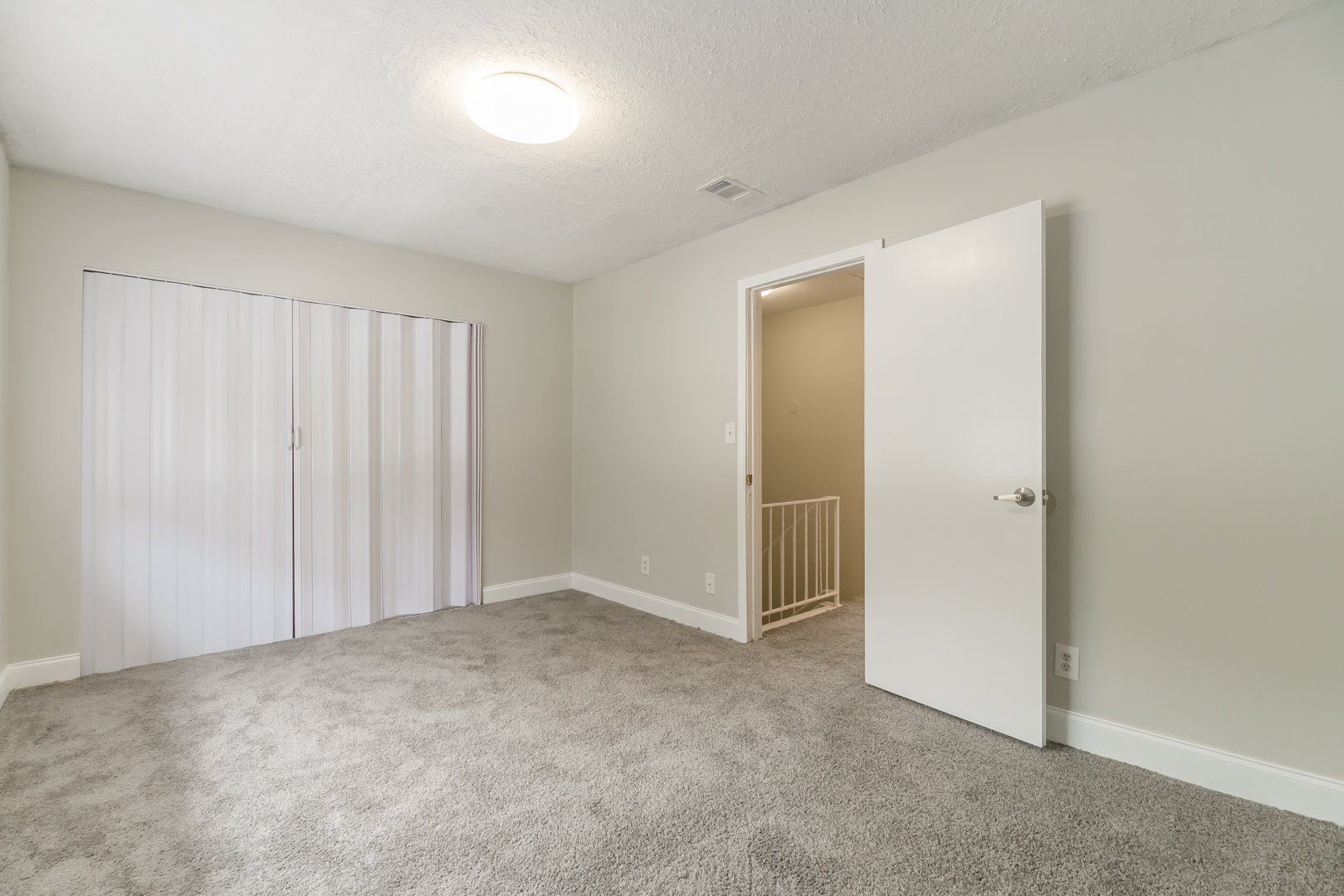 Empty beige bedroom with gray carpet, white closet doors, and an open door to a hallway.