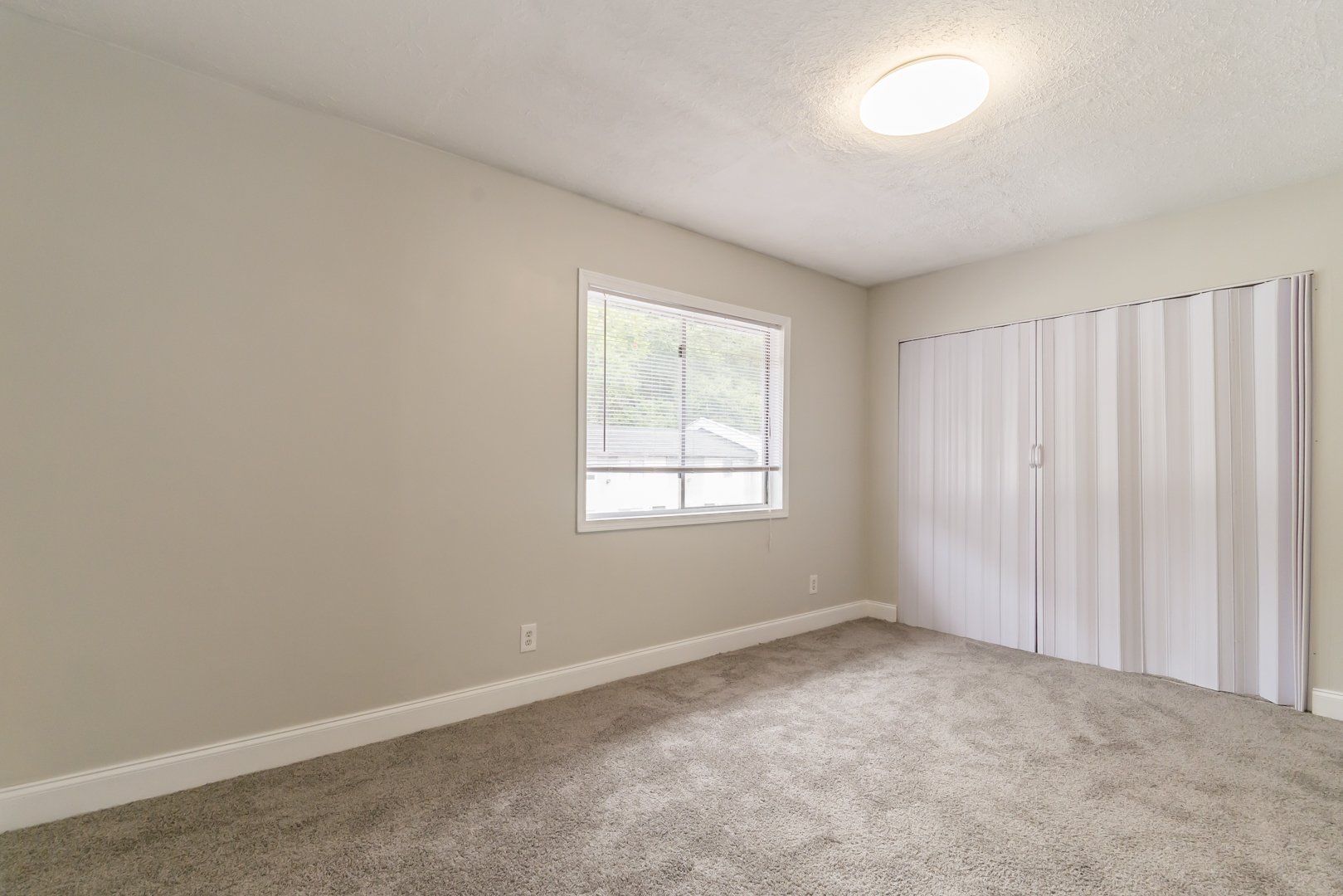 Empty beige bedroom with carpet, window, ceiling light, and white closet doors