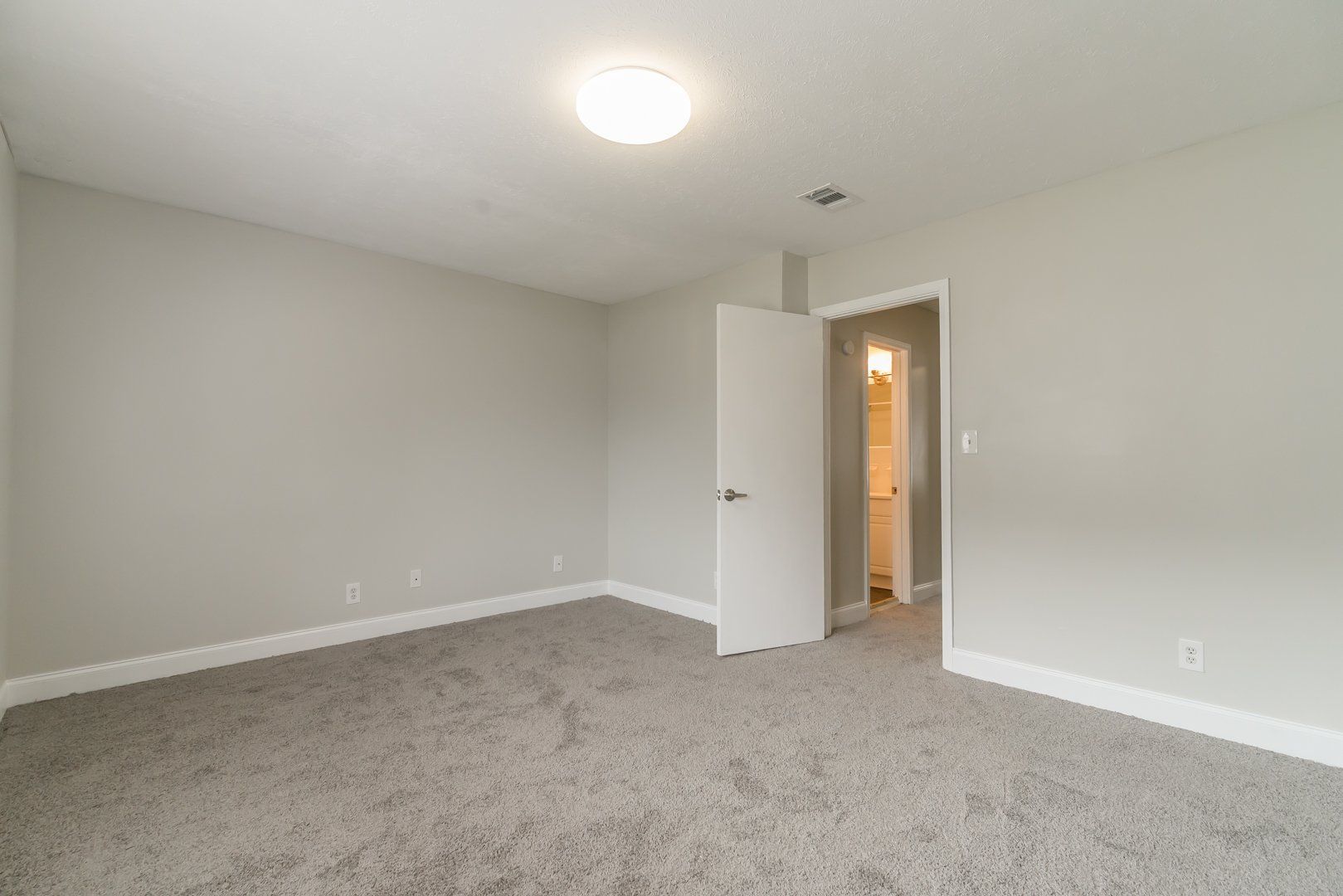 Empty beige bedroom with carpet, white walls, open door, and ceiling light.