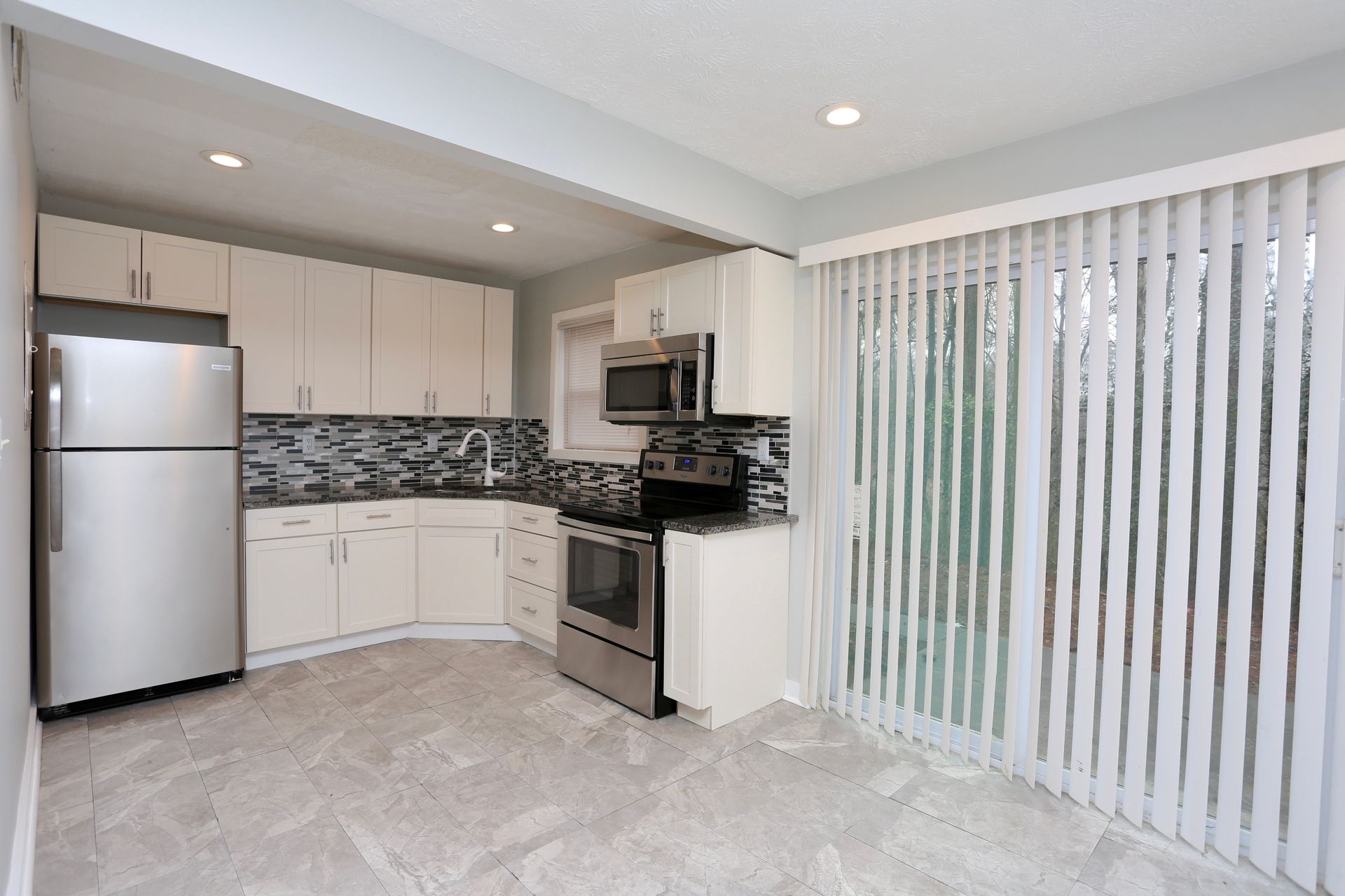 Bright kitchen with white cabinets, stainless fridge, tiled backsplash, and vertical blinds beside a dining area