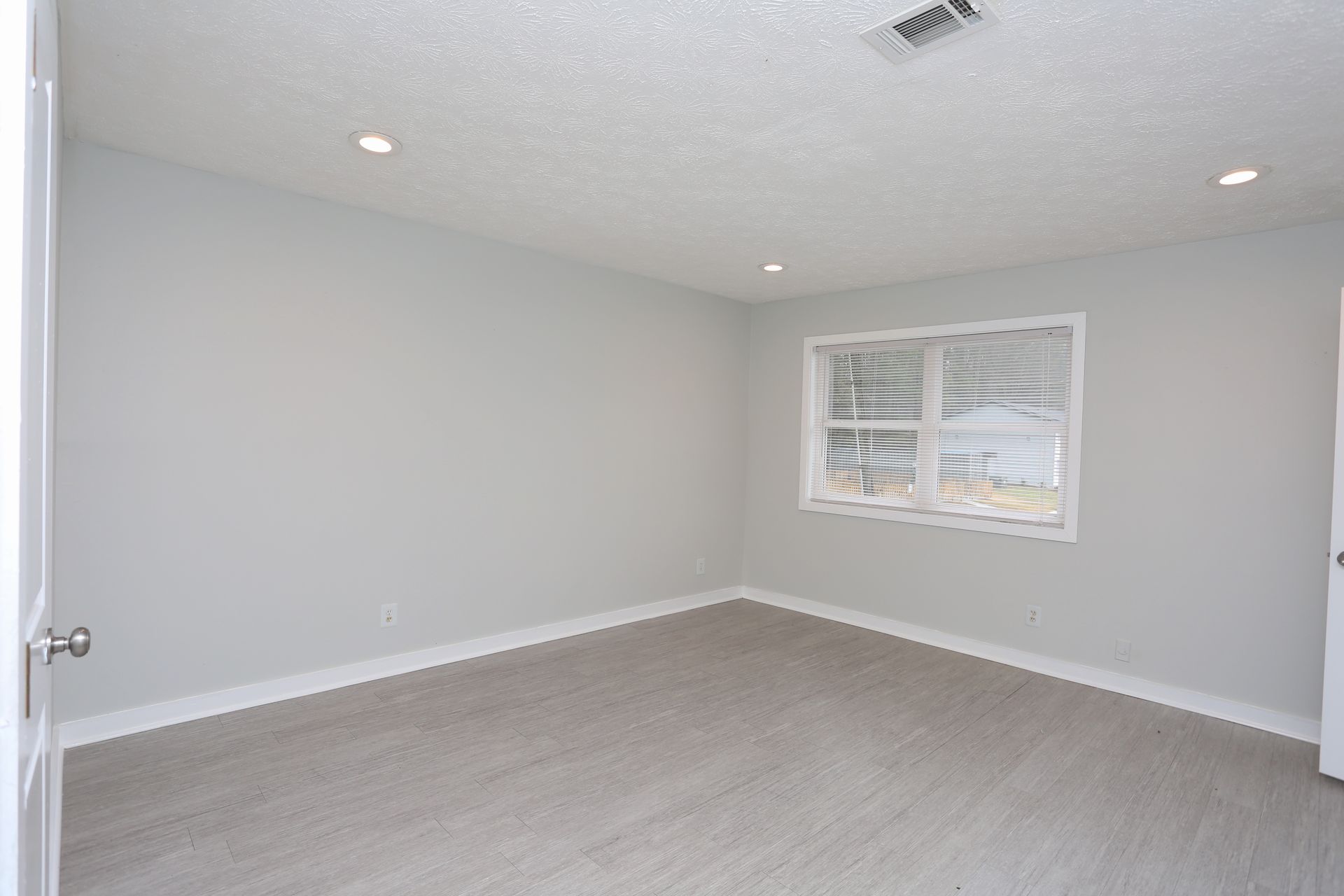 Empty light-gray bedroom with carpet, white walls, recessed lights, and a window with blinds.