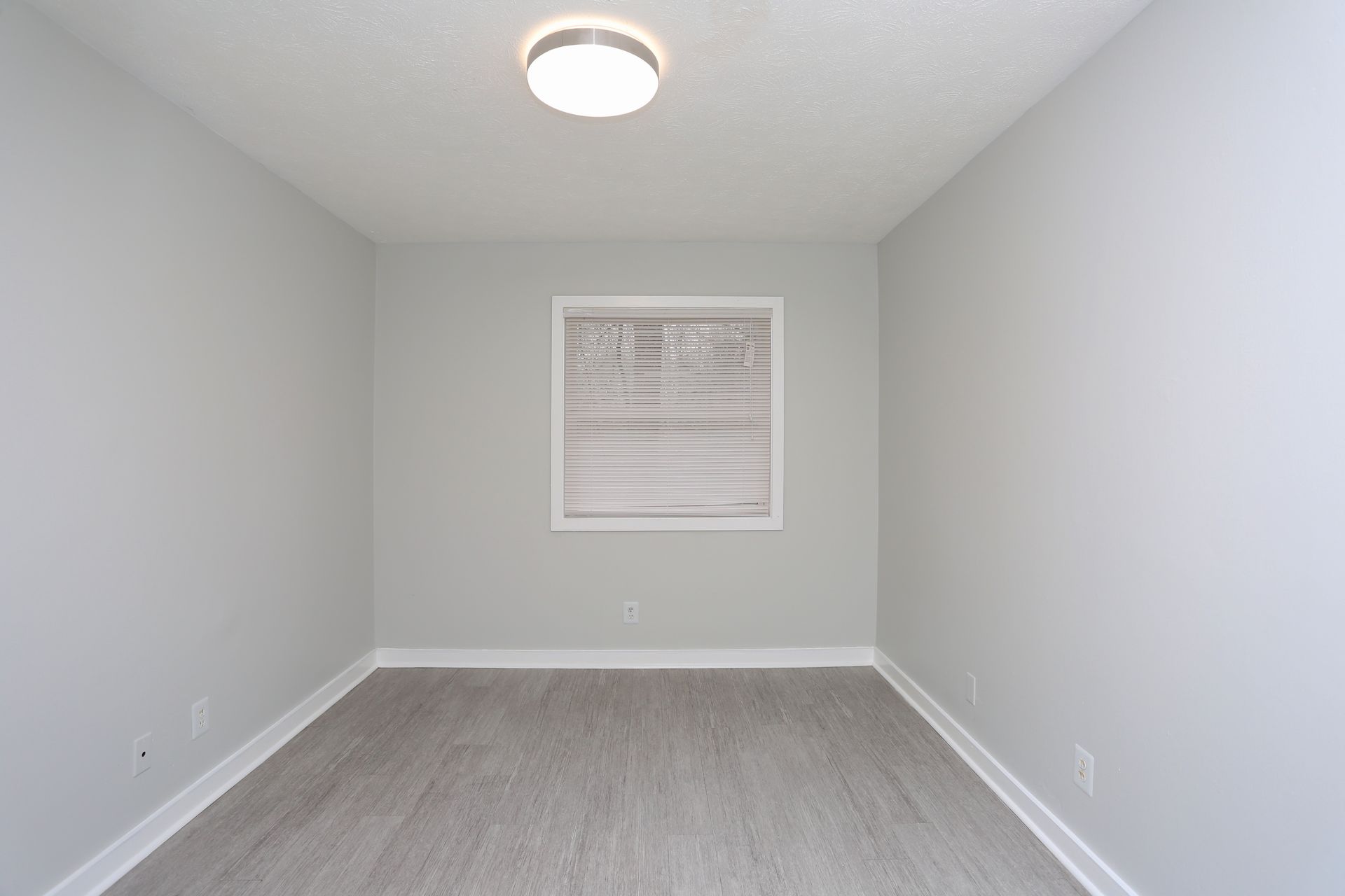 Empty gray room with carpet, a ceiling light, and a small window with blinds.