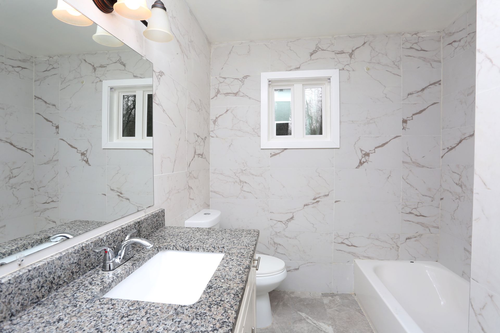 Modern bathroom with marble-look tile, granite vanity, bathtub, and two small windows.