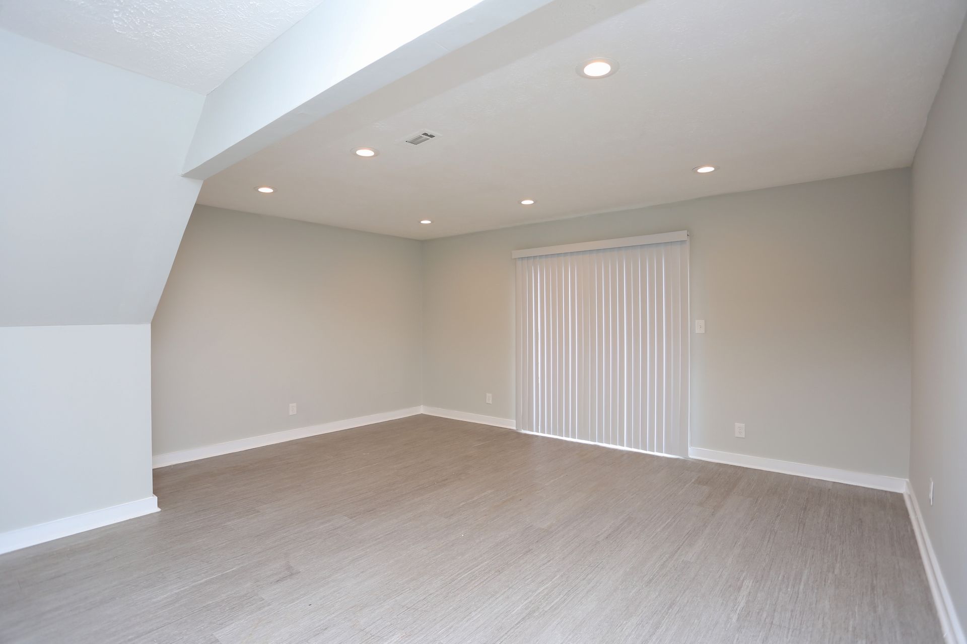 Empty beige carpeted room with vaulted ceiling, recessed lights, and vertical blinds on the far wall