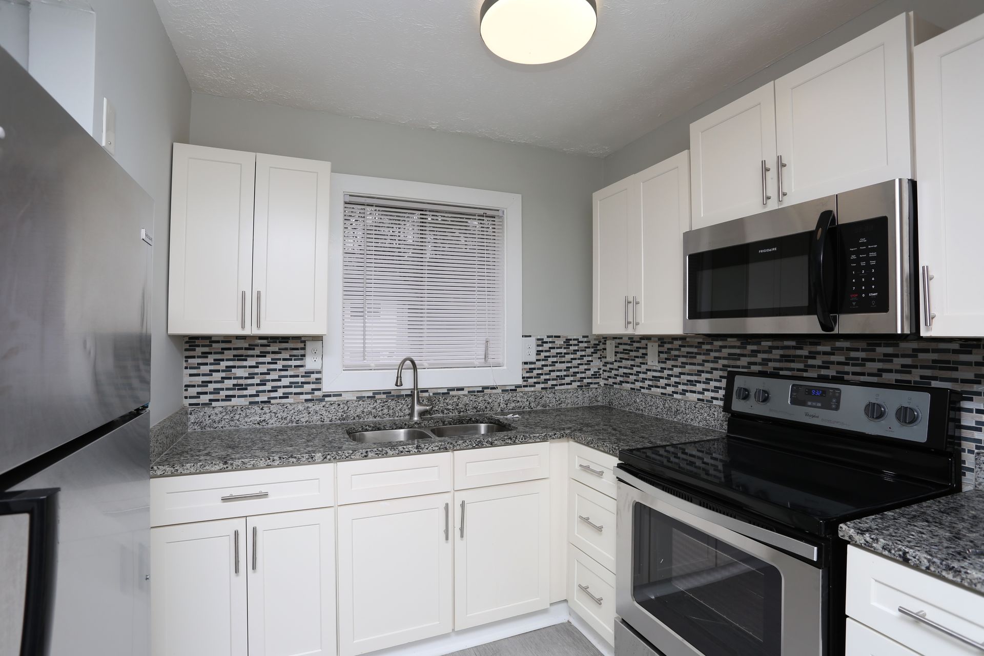 Modern white kitchen with black appliances, gray mosaic backsplash, and window above the sink
