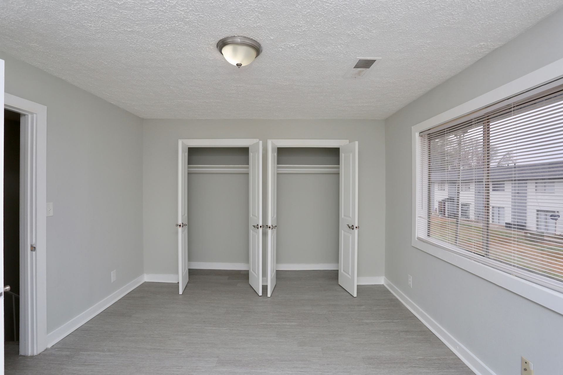 Empty light-gray bedroom with two open closets, white trim, and a window with blinds.