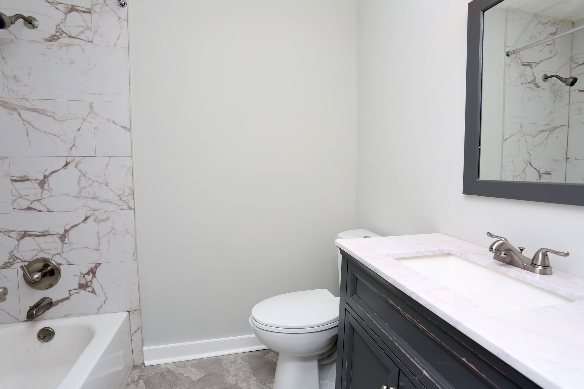 Small modern bathroom with white walls, marble shower tile, toilet, and dark vanity with sink and mirror