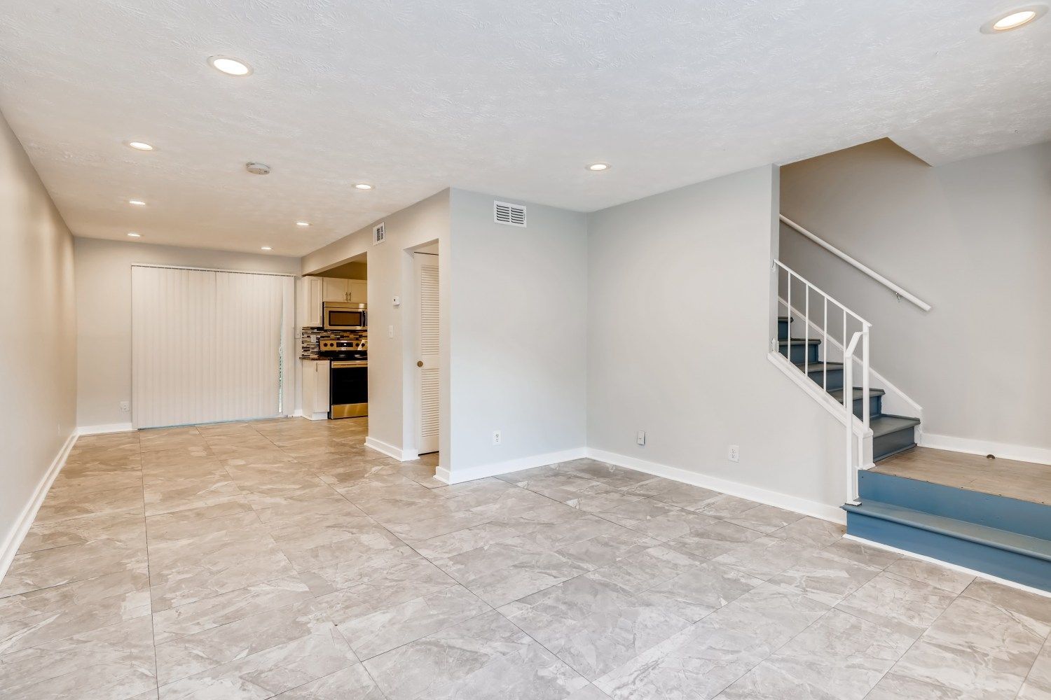 Empty beige living room with tiled floor, recessed lights, and a staircase on the right