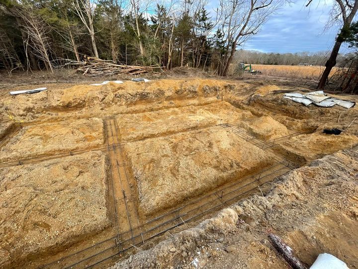 Excavated foundation with rebar laid in trenches, surrounded by dirt and trees.