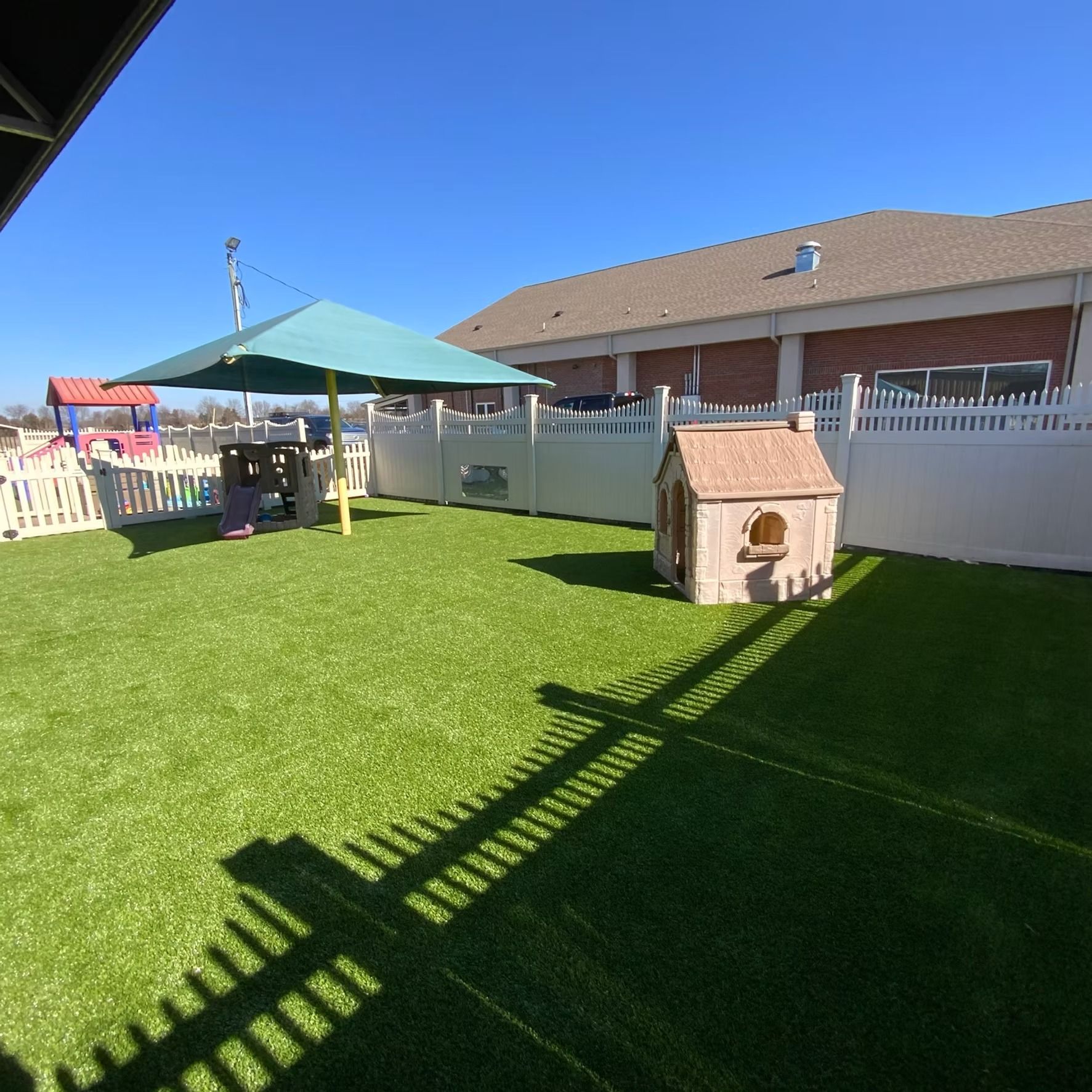 Playground with green turf, white fence, playhouse, and umbrella on a sunny day.