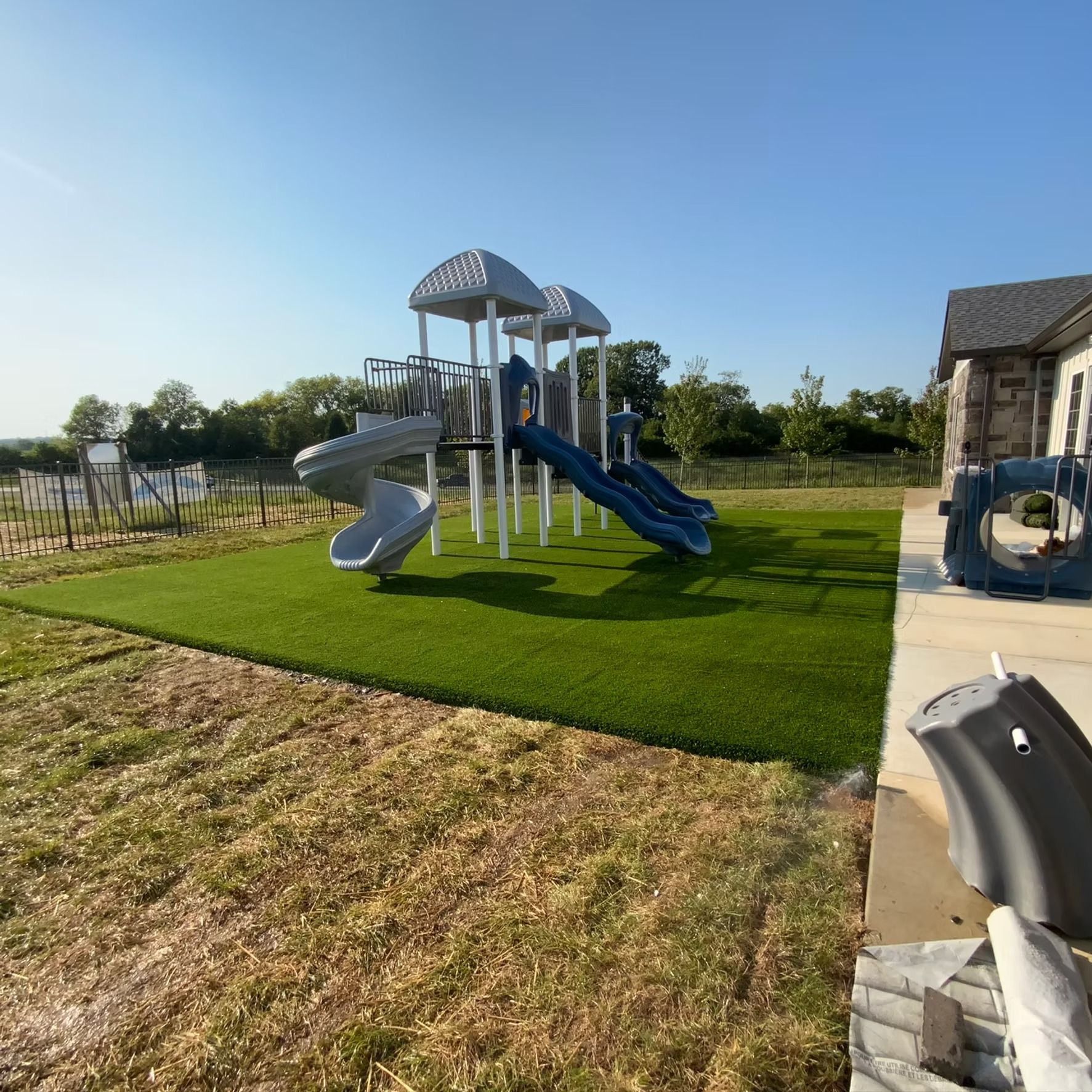 Playground with slides on artificial turf, next to a building and fence, under a blue sky.