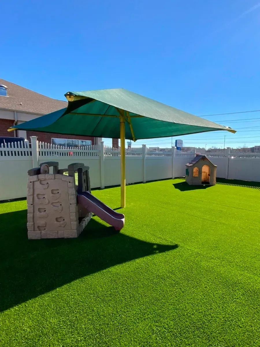 Playground with green shade, slide, playhouse, and artificial grass.