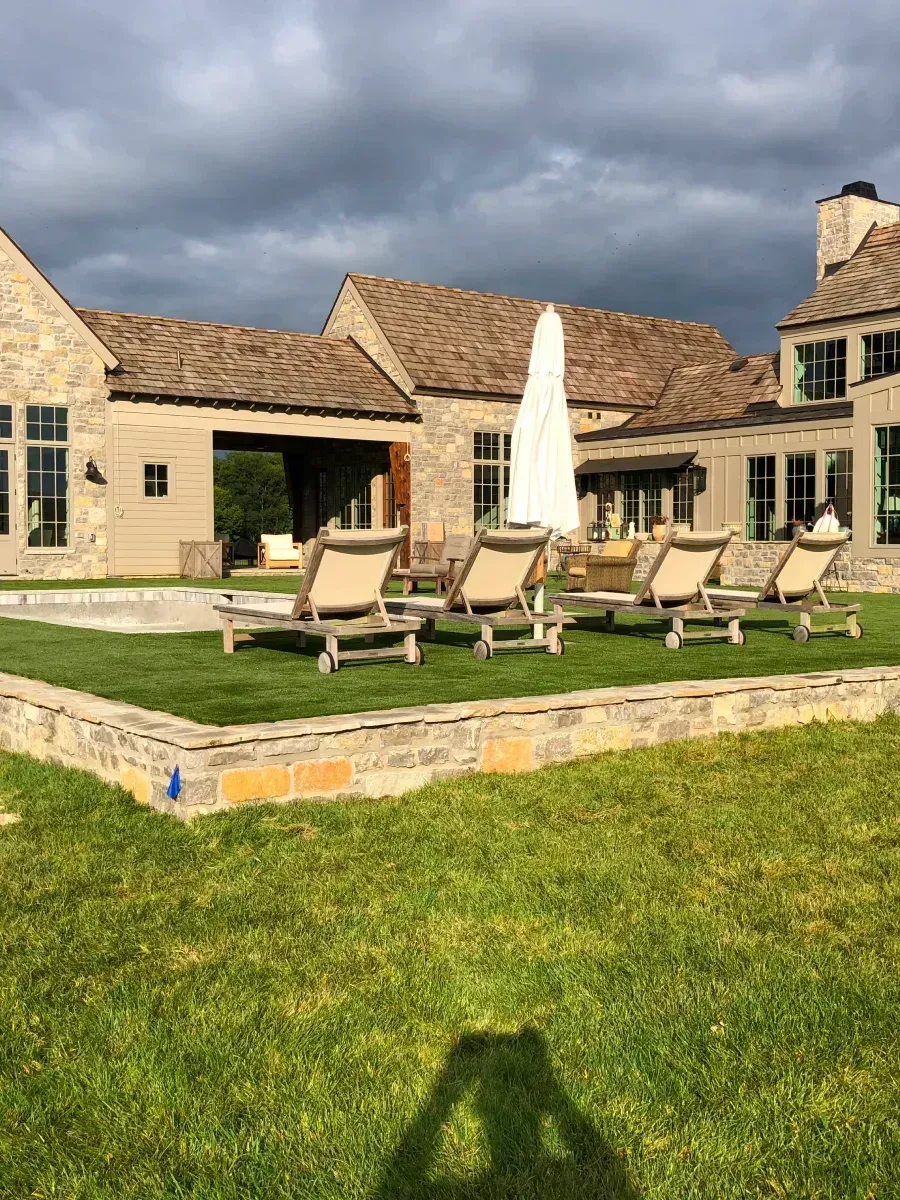 Lounge chairs on a lawn by a pool in front of a stone house under a cloudy sky.