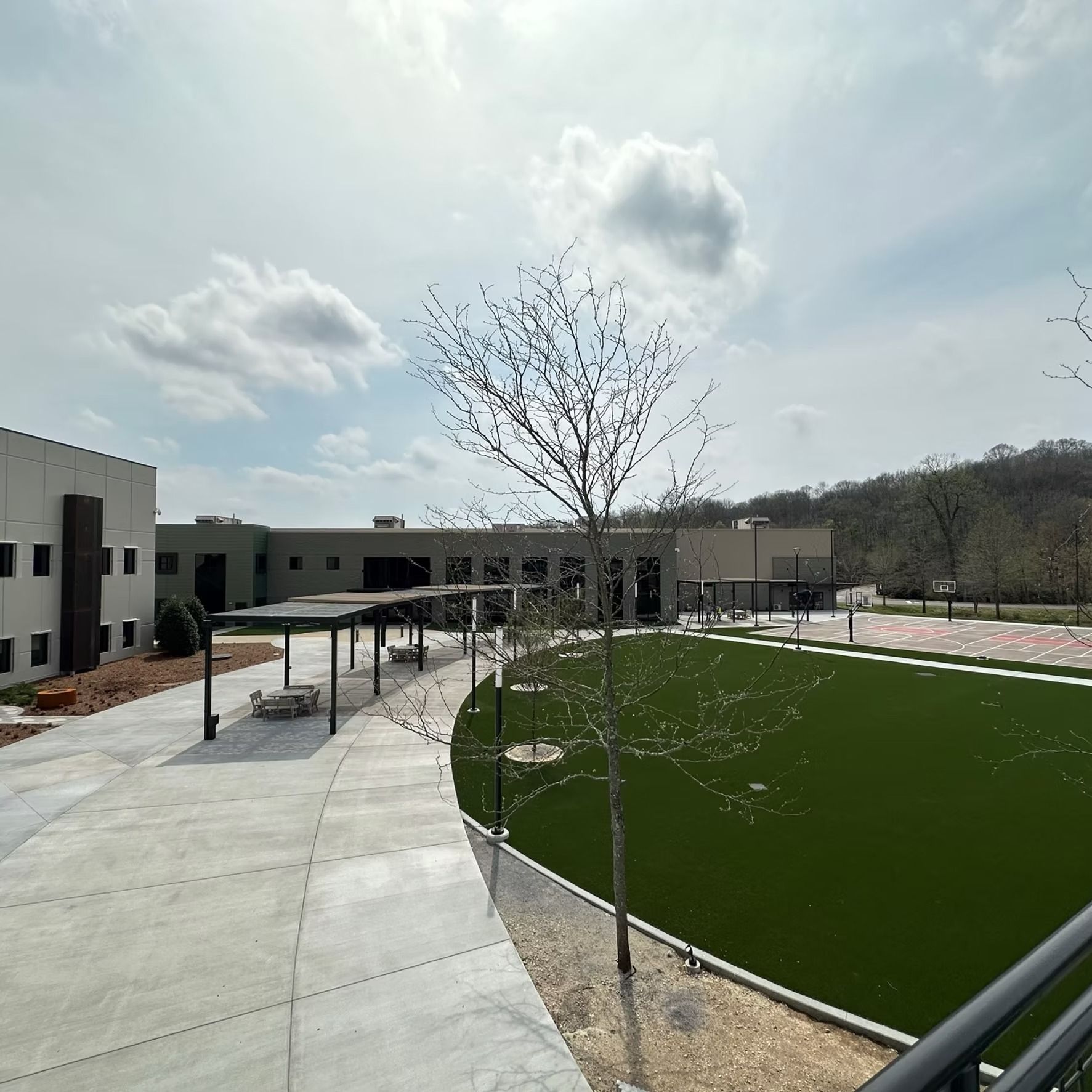 Exterior view of a modern building with a walkway, green space, and a tree, under a cloudy sky.