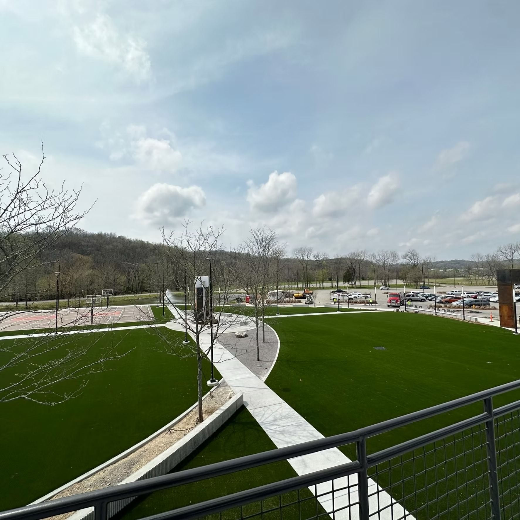 Park with green grass, walkways, trees, and parking area under a cloudy sky.