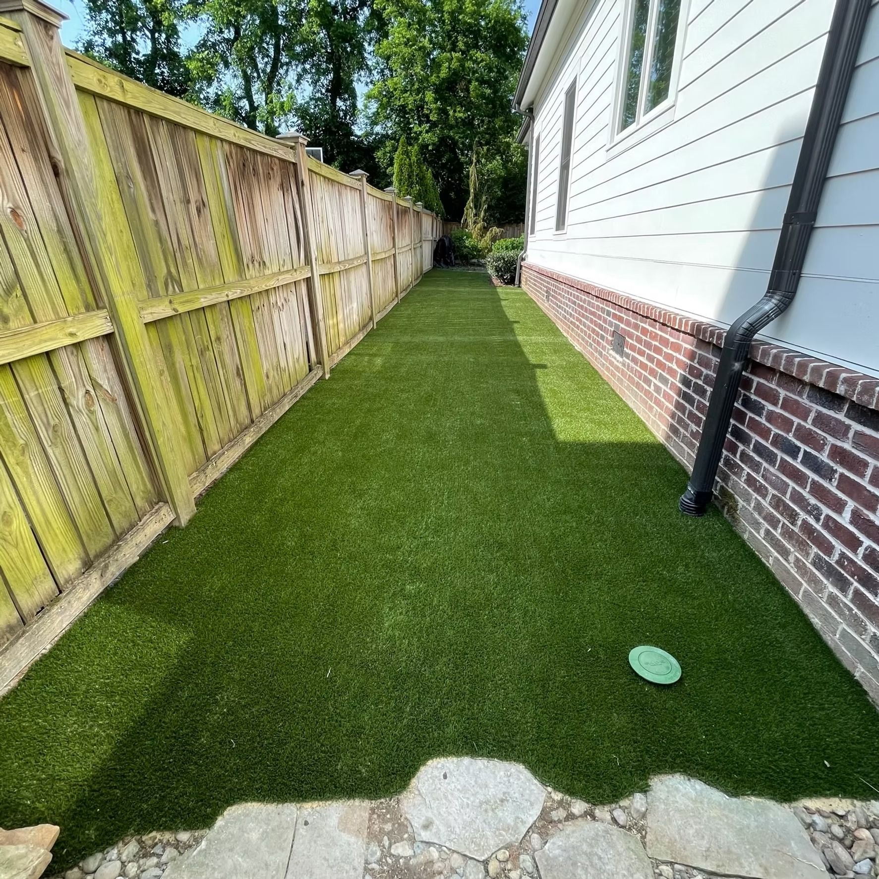 Narrow backyard with artificial turf between a wooden fence and a house with brick foundation.