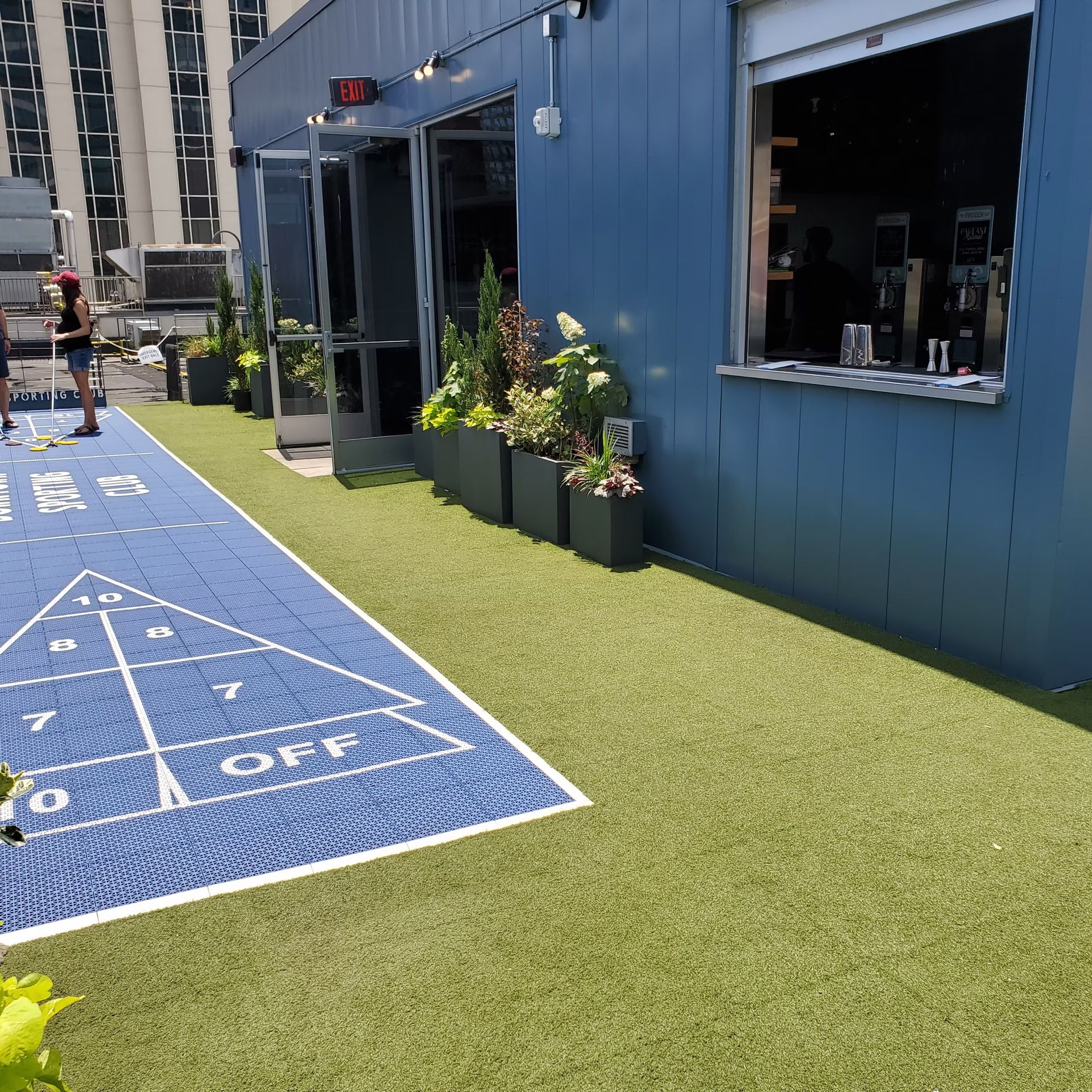 Shuffleboard court on blue surface, green turf, blue building with open windows, potted plants.