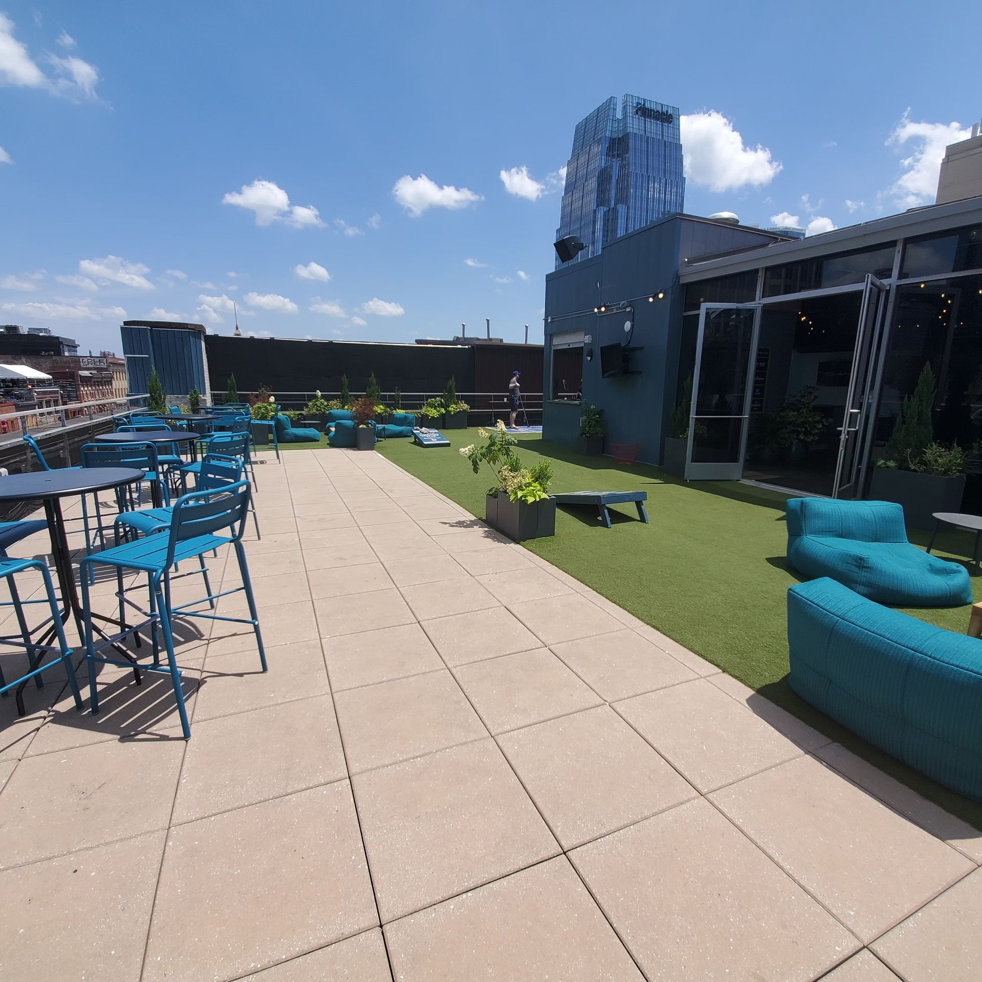 Rooftop patio with blue tables and chairs, turf, and buildings in the background on a sunny day.