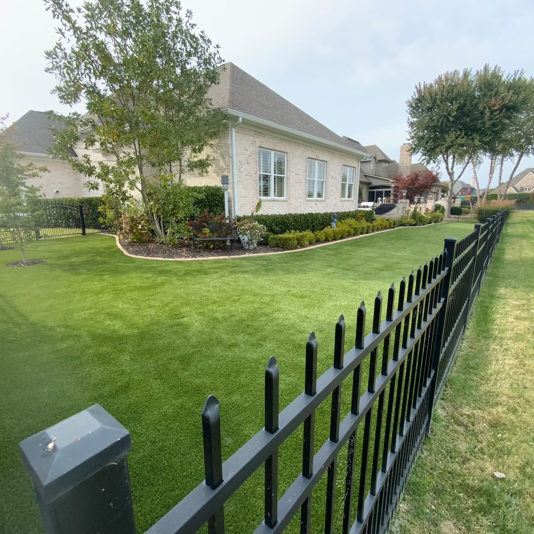 Black metal fence bordering a manicured green lawn along a light brick house on an overcast day.