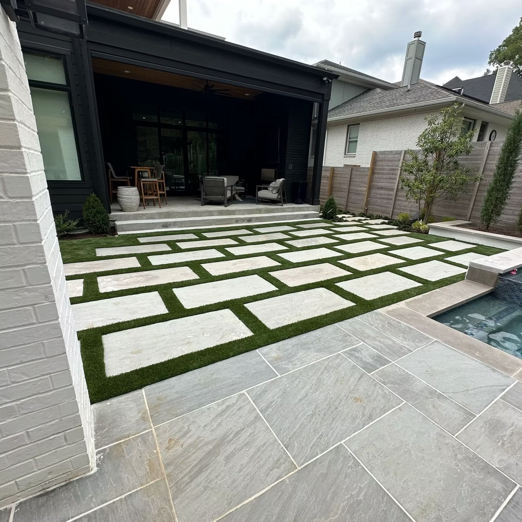 Backyard patio with stone pavers and artificial turf, near a pool and modern house.