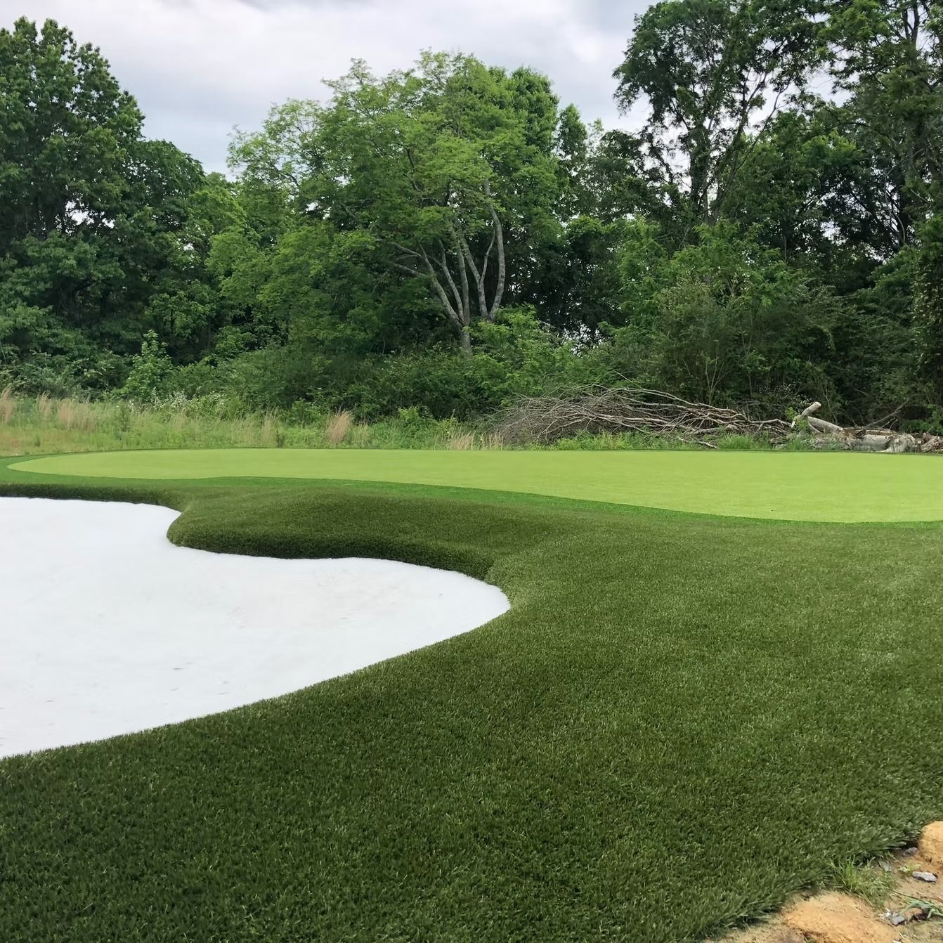 Golf green with artificial turf, white sand trap, and trees in the background.