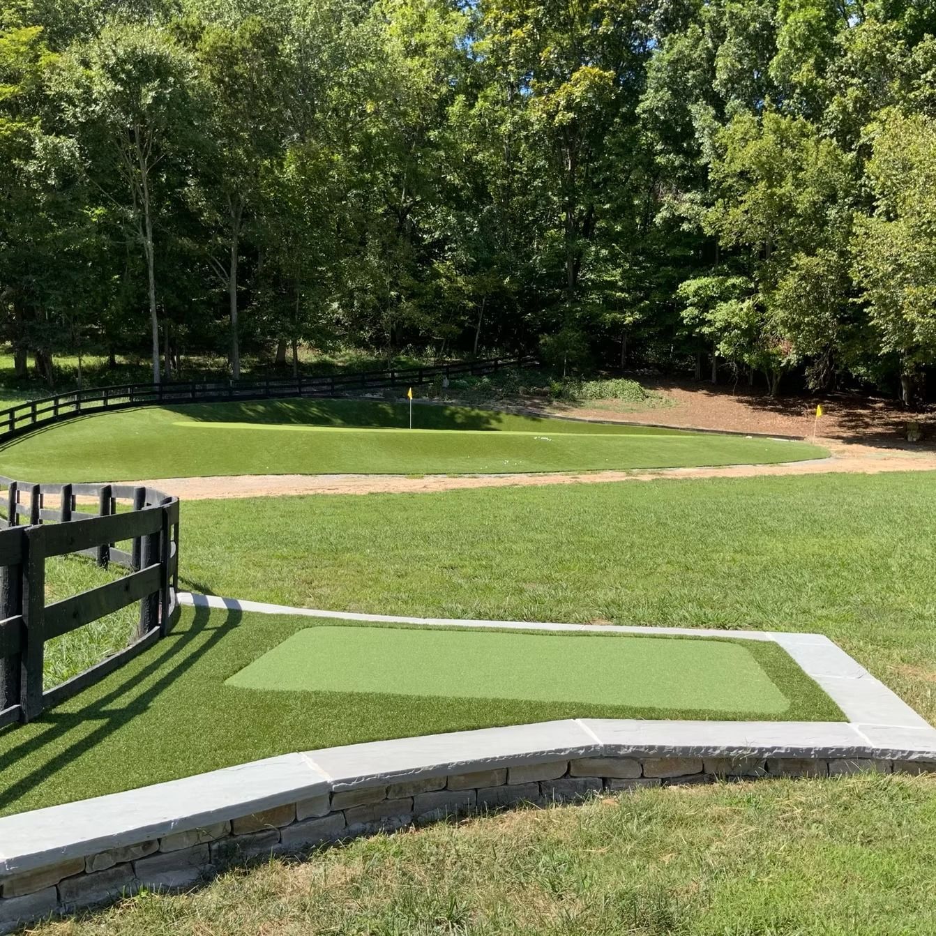 Golf green with putting area surrounded by grass and trees. Black fence on the left.