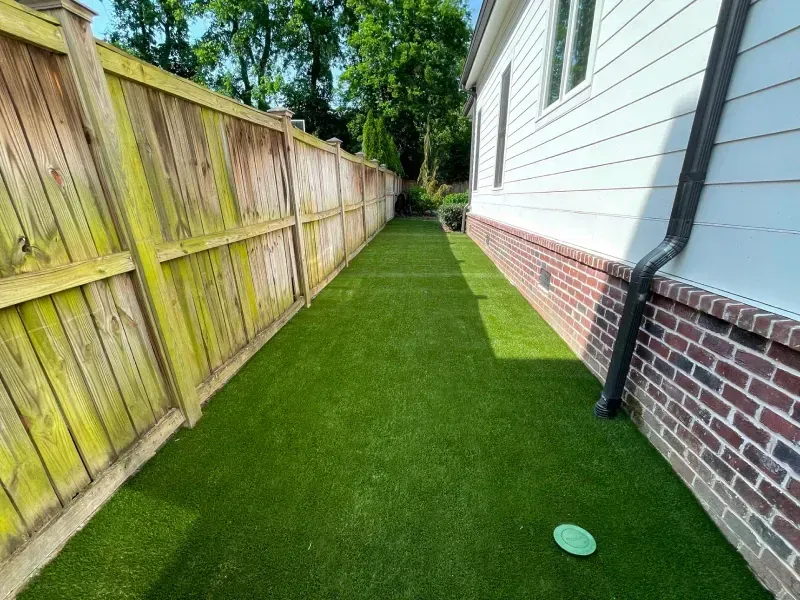 Narrow yard with artificial turf between a wooden fence and a brick-based house.