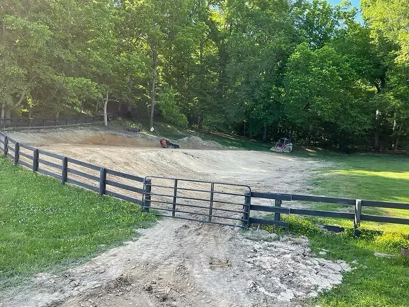 Dirt driveway leads to a construction site behind a black wooden fence; trees in the background.