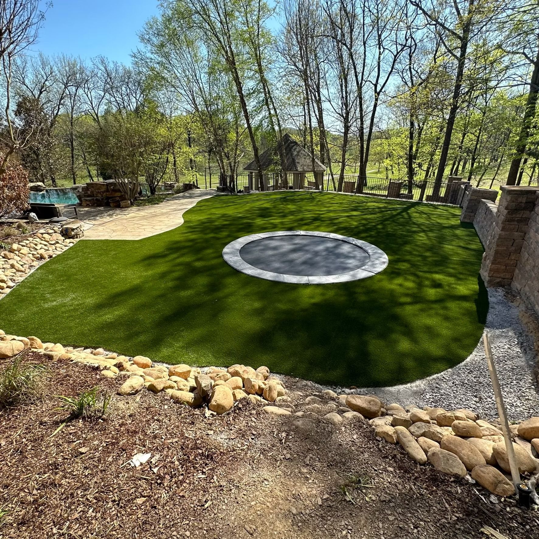Green lawn with a trampoline, surrounded by rocks and trees.