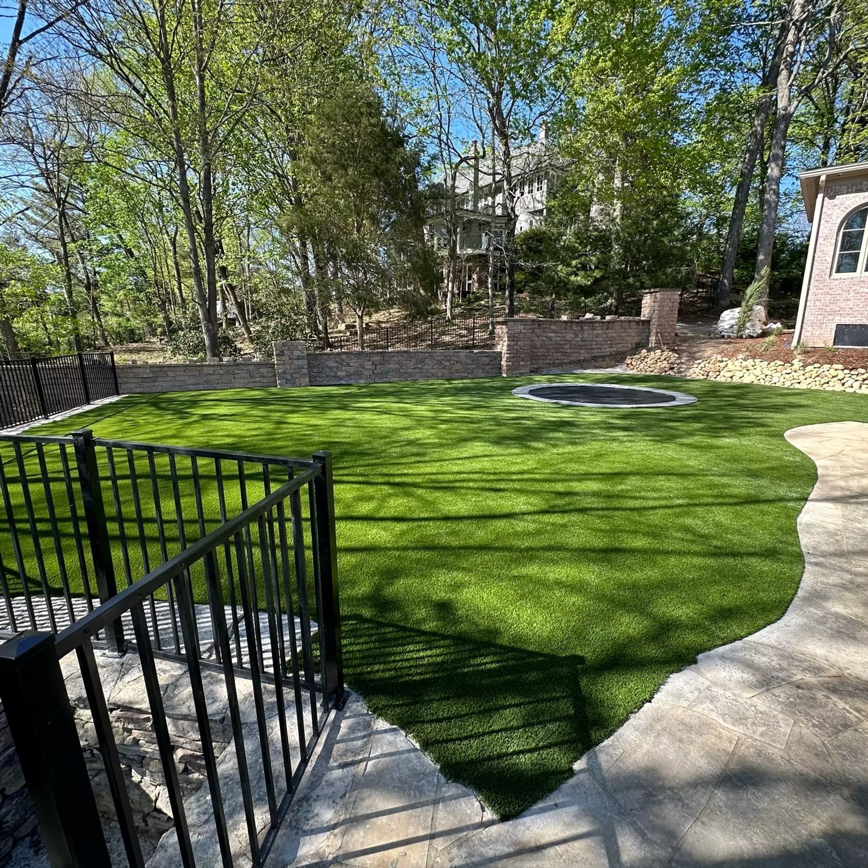 A lush green lawn with a trampoline, surrounded by a brick wall and trees, viewed from stairs.