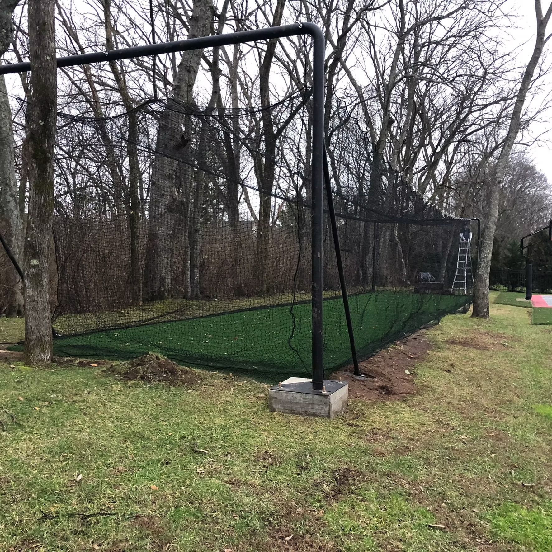 Black netting and metal frame covering a green turf area, in a wooded, outdoor setting.