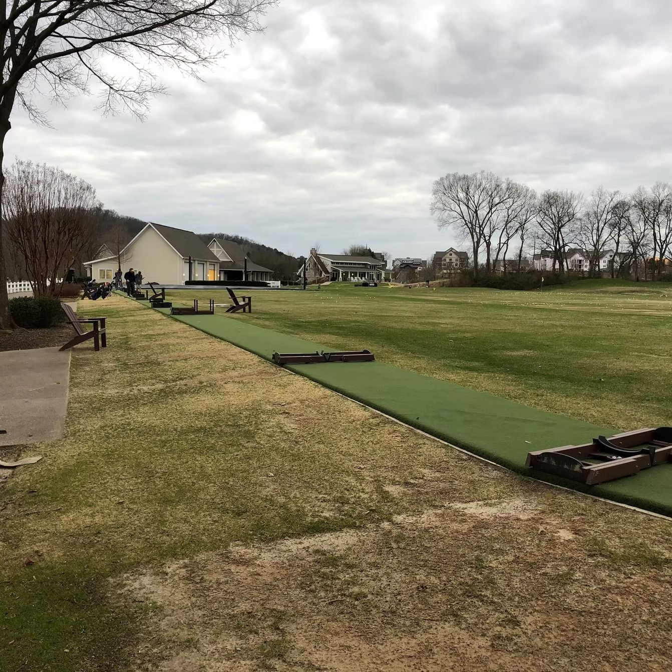 A large grassy area with buildings in the background. A dark green strip of turf runs across the ground. Overcast day.
