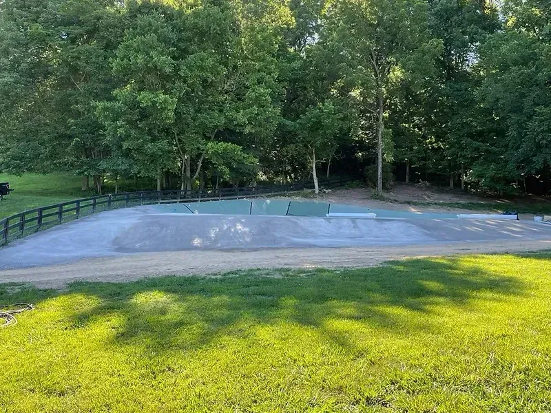 A concrete skatepark with green and gray features, set in a grassy park, bordered by trees.
