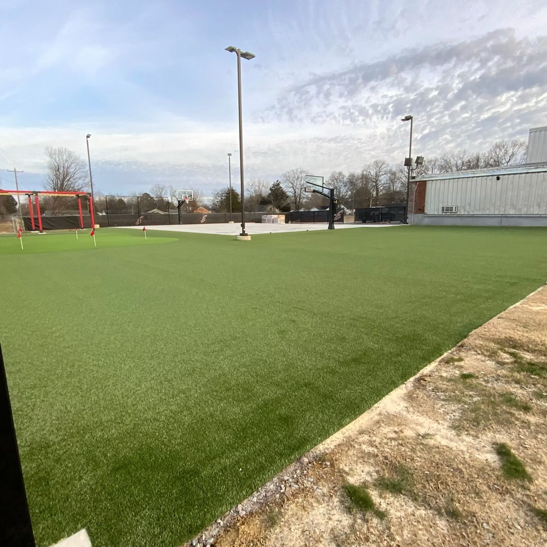 Artificial turf sports field with basketball hoop and soccer goal under cloudy sky.