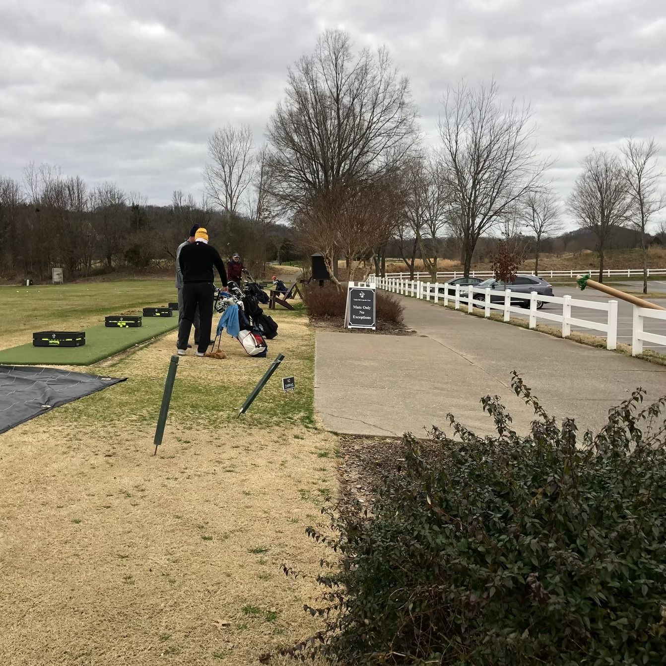 Golf driving range on a cloudy day. Several people near golf bags and mats. Path with white fence.