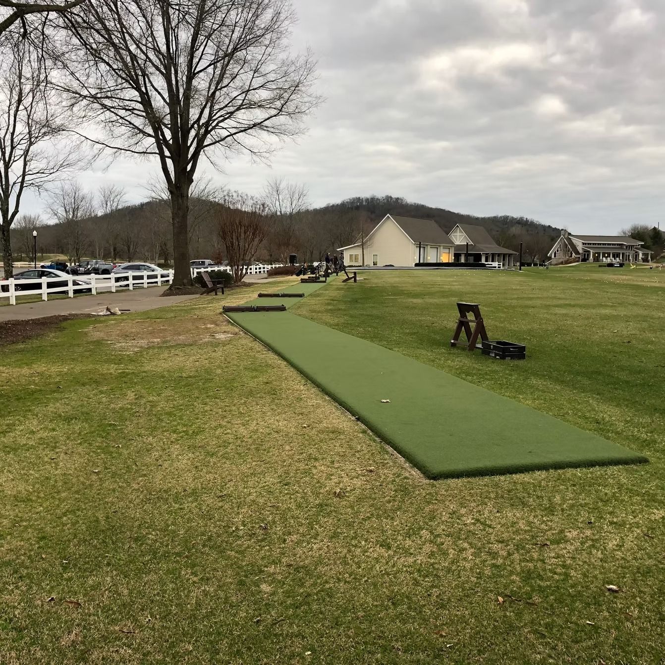 Artificial putting green on a grassy area, with a building and parked cars in the background. Overcast sky.