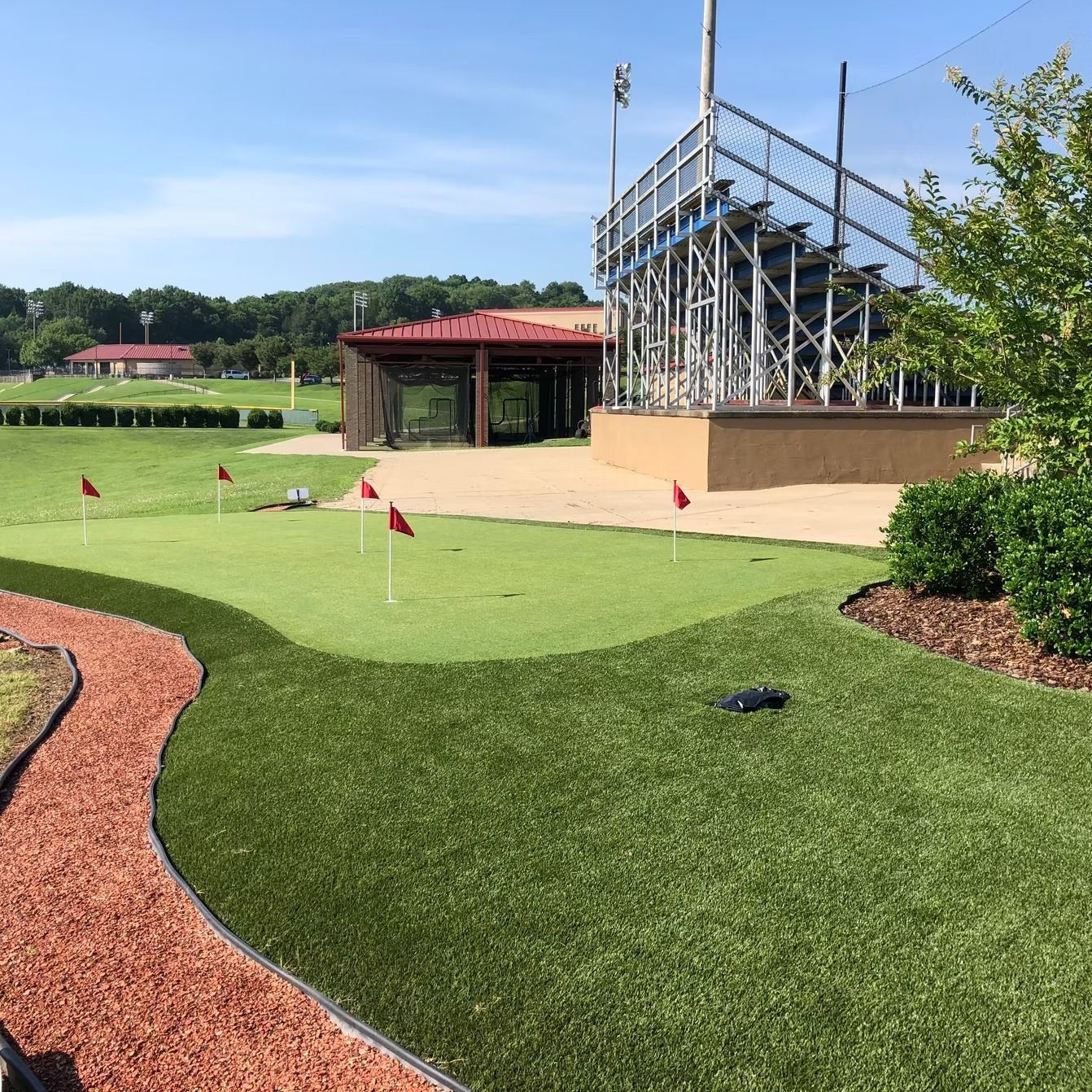 Miniature golf course with green turf, red flags, pathway, and bleachers on a sunny day.
