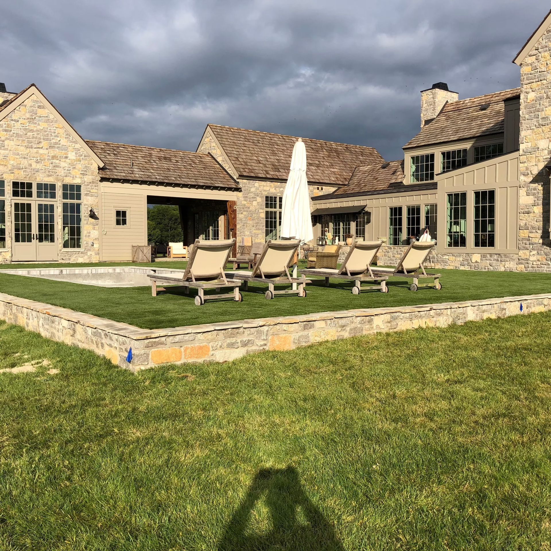Lounge chairs near a pool on a grassy lawn, in front of a large stone house under a cloudy sky.