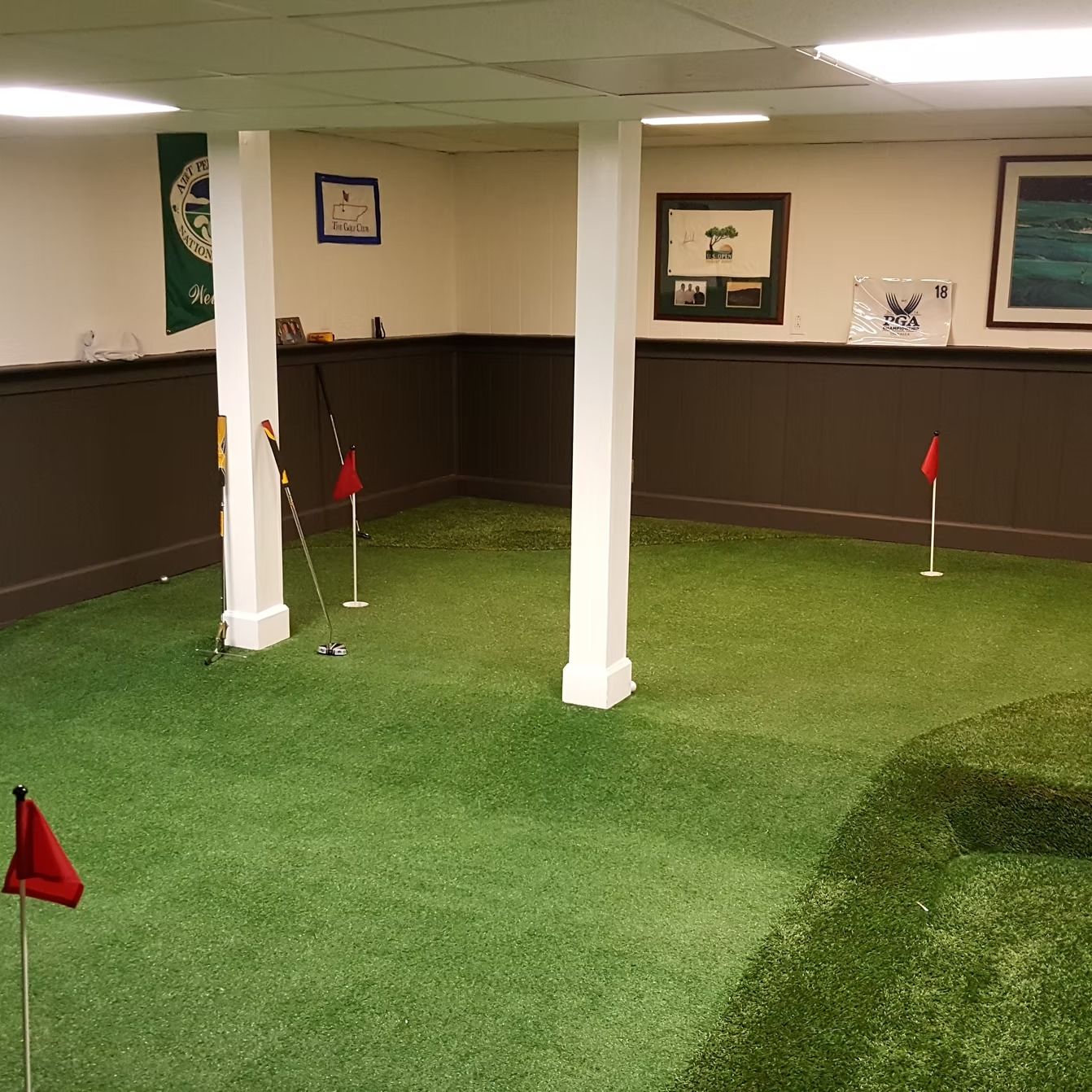 Indoor putting green with golf flags and clubs. Room has white columns, dark brown paneling, and framed art.