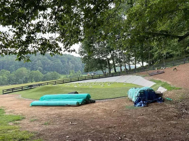 A green field with construction materials, a wooden fence, and trees in the background under an overcast sky.