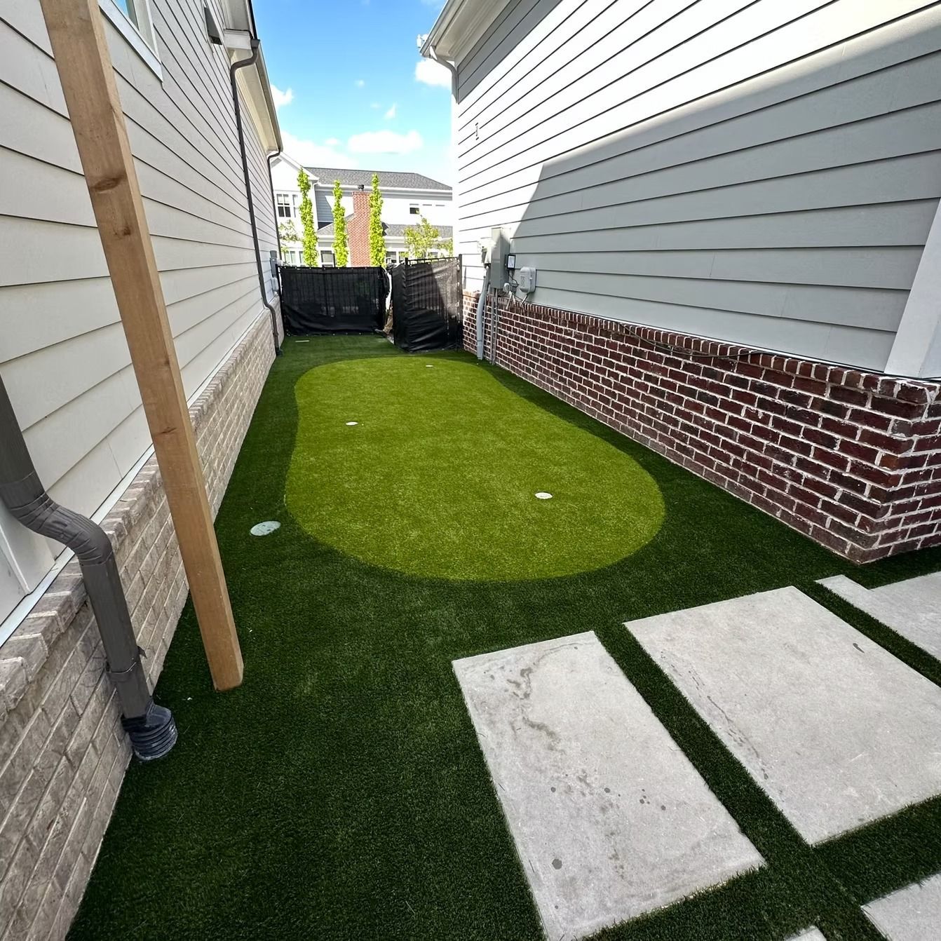 Narrow backyard with artificial turf, bordered by brick and siding, concrete stepping stones.
