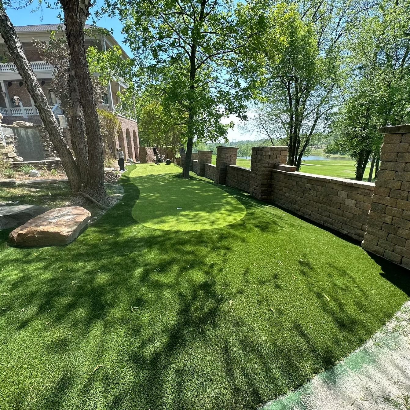 A green lawn with a stone wall and building. Trees cast shadows. Sunny outdoor scene.