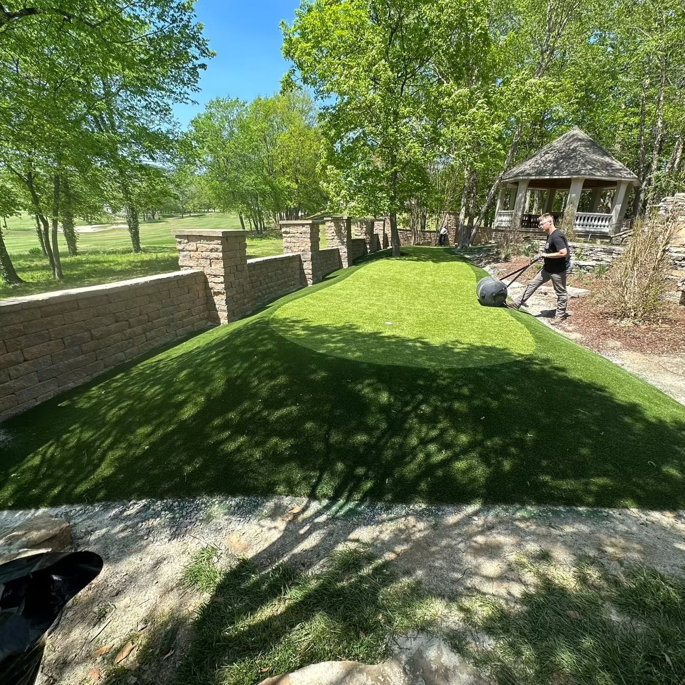 Man rolling turf on a putting green, next to a stone wall and gazebo, under a sunny sky.